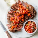 Raw flank steak being prepared with garlic and olive oil on a marble countertop.